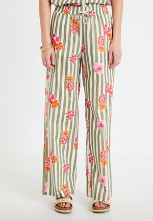 Luźne spodnie z pionowymi zielono-białymi paskami, ozdobione różowymi, czerwonymi i pomarańczowymi ananasami oraz kwiatami, noszone z beżowymi sandałami.