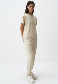Beige top met korte mouwen en een horizontale naad, gecombineerd met bijpassende tapered broeken. Glad materiaal, minimalistisch ontwerp en witte sneakers.
