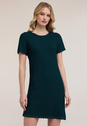 Femme blonde portant une robe en maille vert foncé à manches courtes avec des bords festonnés, se tenant devant un fond clair uni.