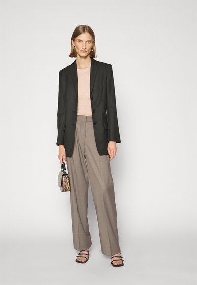 Zwarte op maat gemaakte blazer over een lichtroze top, gecombineerd met beige brede broekspijpen en zwarte sandalen met bandjes, terwijl ze een kleine met een patroon versierde handtas vasthoudt.