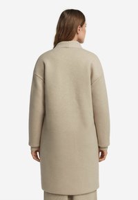 Capotto lungo beige realizzato in materiale morbido, con un taglio dritto, spalle cadenti e dettagli a coste sui polsini. Design semplice e minimale.