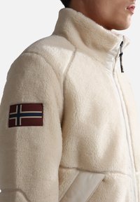 Fleece jacket in off-white with a high collar, featuring a textured surface and a maroon patch with a white and blue flag on the sleeve.