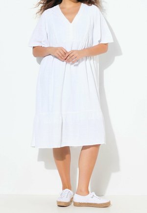 Femme portant une robe blanche ample mi-longue aux genoux avec manches courtes et des baskets blanches à semelles tissées sur fond blanc.