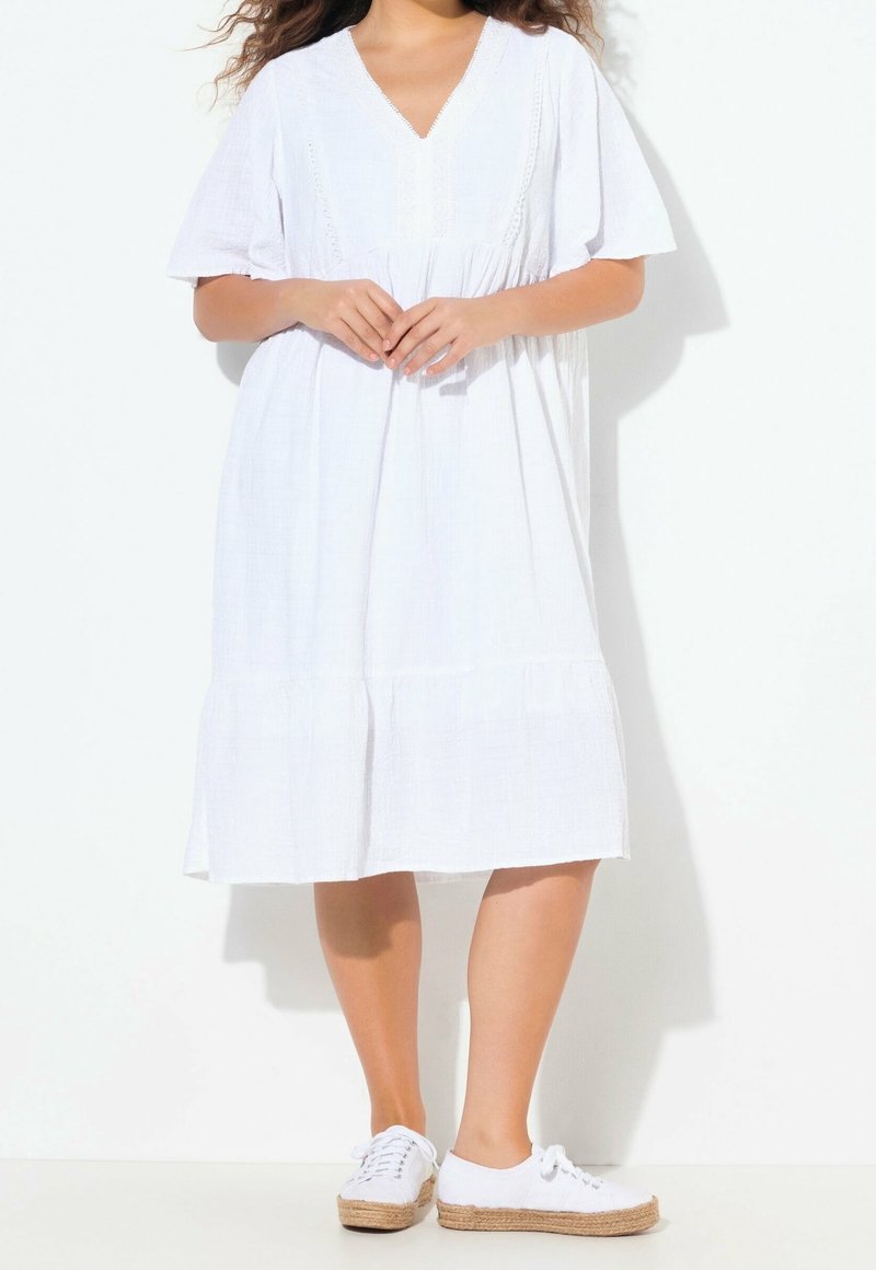 Femme portant une robe blanche ample mi-longue aux genoux avec manches courtes et des baskets blanches à semelles tissées sur fond blanc.