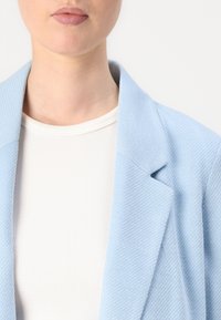 Απαλό μπλε υφασμάτινο σακάκι με εσοχές, φορεμένο πάνω από ένα λευκό ριγωτό τοπ. Εστίαση στη λεπτομέρεια του υφάσματος και την αντίθεση χρώματος.