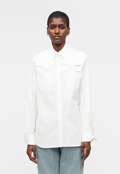 Femme portant une chemise blanche boutonnée avec col pointu et poches à rabat, associée à un jean bleu clair, debout devant un fond uni.