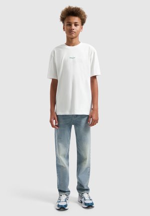 Tienerjongen staand, draagt een wit T-shirt met korte mouwen, lichtblauwe spijkerbroek en witte en blauwe sneakers.