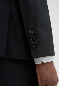 Dunkel gemusterte Anzugjacke mit einreihigem Design. Sie verfügt über eine weiße Hemdmanschette mit abgeschrägten Kanten und vier schwarzen Knöpfen am Ärmel.