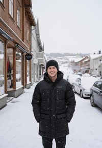 Czarna kurtka puchowa z teksturowanym kapturem, wyposażona w dwie przednie kieszenie i zapięcie na zamek, noszona na czarnych spodniach na zaśnieżonej ulicy.