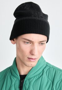 Bonnet en tricot noir avec une texture côtelée. Porté de manière décontractée, il présente un sommet lisse et un subtil motif géométrique. Accompagné d'une veste matelassée verte.