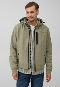 Veste softshell vert olive avec capuche, dotée d'une fermeture éclair, d'accents noirs et de deux poches avant ; portée sur une chemise à carreaux.