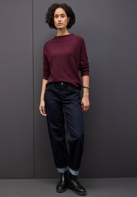 Burgunderfarbener Strickpullover, dunkle Jeans mit aufgerollten Säumen und schwarze Stiefeletten. Das Outfit zeichnet sich durch einen lockeren Schnitt und ein einfaches Design aus.