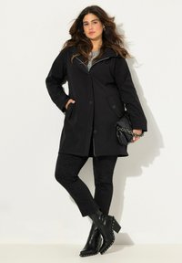 Cappotto nero fino al ginocchio con chiusura a zip e bottoni, dotato di tasche laterali, abbinato a pantaloni neri con motivo e stivaletti con borchie.