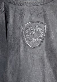 Giacca in pelle grigia con superficie texturizzata, dettagli cuciti e logo ricamato a forma di scudo con design a corona.