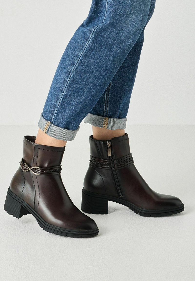 Personne portant des bottines en cuir marron foncé avec des fermetures éclair latérales et un détail tressé, associées à un jean bleu retroussé.
