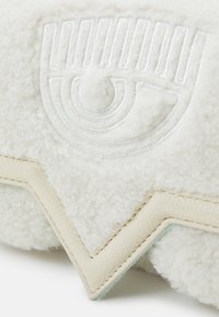 Biała pluszowa poduszka z wytłoczonym wzorem oka. Teksturowana, miękka wełniana wykończenie, uzupełniona jasnobeżowym skórzanym akcentem wzdłuż krawędzi.