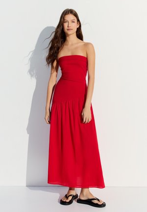 Junge Frau mit langen braunen Haaren, die ein trägerloses rotes Maxikleid und schwarze Flip-Flops trägt, steht vor einer weißen Wand.
