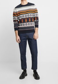 Marinblå stickad tröja med flerfärgade geometriska och festliga mönster, rund halsringning och ribbade muddar; kombinerad med mörkblå jeans och svarta skor.