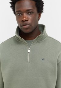 Mężczyzna z krótkimi, kręconymi włosami, ubrany w jasnozieloną bluzę na zamek z małym logo konia na lewej piersi, na jednolitym tle.