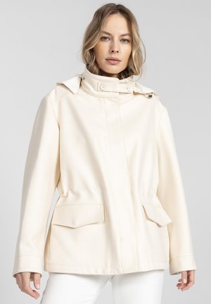 Kobieta w kremowej kurtce z wysokim kołnierzem i ozdobnymi guzikami, stojąca na tle jednolitego jasnoszarego tła.