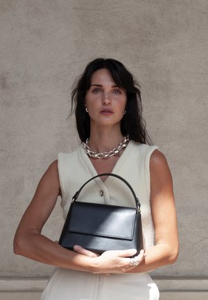 Mujer de cabello oscuro con una blusa sin mangas beige y un collar plateado, sosteniendo un bolso de cuero negro frente a una pared lisa.