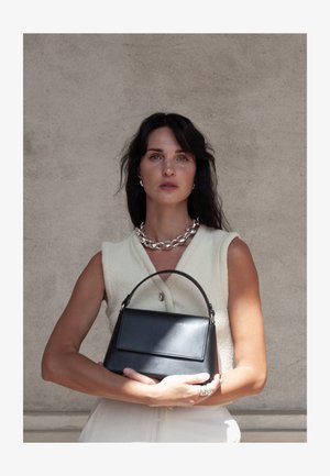 Γυναίκα με σκούρα μαλλιά που φορεί αμάνικη μπεζ μπλούζα και ασημένιο κολιέ, κρατώντας μια μαύρη δερμάτινη τσάντα μπροστά σε απλή τοίχο.