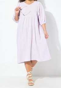 Helles Lavendelkleid in Midi-Länge aus strukturiertem Stoff, mit V-Ausschnitt, gerafftem Bund und Dreiviertelärmeln; kombiniert mit weißen, schnurgebundenen Sandalen.