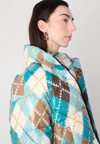 Puffet jakke med et farverigt argyle mønster i teal, brun og creme, med høj krave og lynlås lukning. Glat, blank stof tekstur.