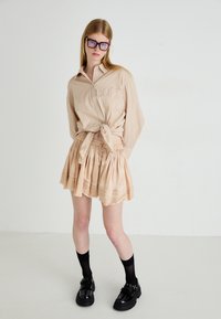 Lichtbruin geruit knoopshirt geknoopt in de taille, gecombineerd met een vloeiende beige geplooide rok, zwarte enkelsokken en stevige zwarte schoenen.