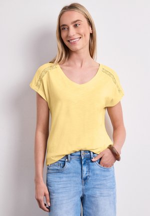 Smilende kvinde stående iført en gul kortærmet top med blonder på skuldrene, lyseblå jeans og en brun armbånd på hendes højre håndled.