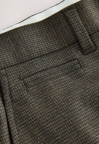 Gros plan sur un tissu texturé gris foncé montrant une ceinture, une boucle de ceinture, et une couture horizontale de la poche arrière sur un pantalon sur mesure.