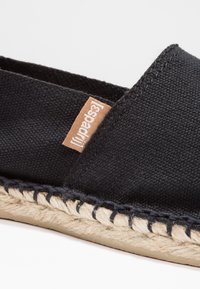 Black canvas espadrille shoe with a jute rope sole and a small brown logo tag. Features stitched detailing along the sole's edge.