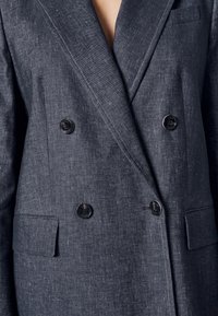Blazer gris foncé à double boutonnage avec revers à pointe, quatre boutons noirs et poches à rabat, porté par une personne.
