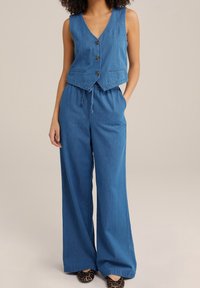 Žena v modré bezrukávové košili s knoflíky a širokých kalhotách na stažení se zvířecím potiskem proti béžovému pozadí.