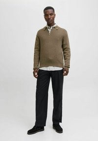 Olive roheline kootud sviiter, millel on luku abil kaelus, on kihiliselt kantud valge ja rohelise triibulise särgi peal, koos mustade kohandatud pükste ja mustade kingadega.