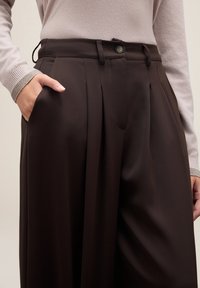 Brązowe, plisowane spodnie z zapięciem na guzik i bocznymi kieszeniami, charakteryzujące się gładką teksturą materiału i dopasowanym krojem.