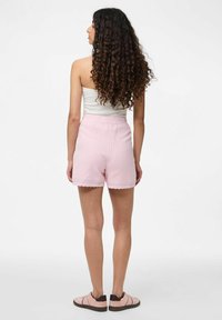 Rozā teksturēti šorti ar viļņotu apakšmali un elastīgu vidukli, kopā ar rozām un brūnām kedām. Aizmugures skats ar redzamām tumšām cirtainām matiņām.