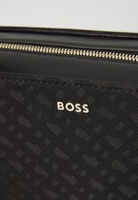 Borsa in tessuto nero con un motivo testurizzato, logo dorato "BOSS" e dettaglio della zip argentata lungo il bordo.