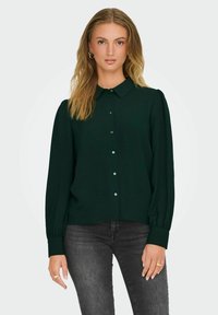 Dunkelgrüne Bluse mit kragenförmigem Ausschnitt, Knopfverschluss und langen Puffärmeln. Aus strukturiertem Stoff mit dezentem Glanz gefertigt.