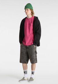 Schwarze Zip-Hoodie, pinkes T-Shirt mit Fronttasche, graue Denim-Shorts, karierten Slip-On-Schuhe und grüne Strickmütze.