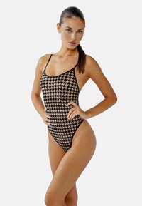 Svart och beige houndstooth-mönstrad baddräkt med tunna axelband och design med hög skärning på benen, tillverkad av ett jämnt, elastiskt tyg.