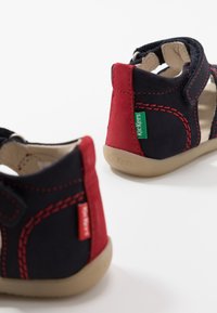 Paire de sandales pour tout-petits bleu marine et rouge avec semelles beige, coutures rouges et étiquettes vertes de la marque Kickers sur les côtés.