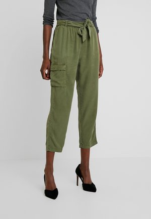 Woman wearing olive green cropped cargo pants with a tied waist and black high-heeled shoes, standing against a plain background.