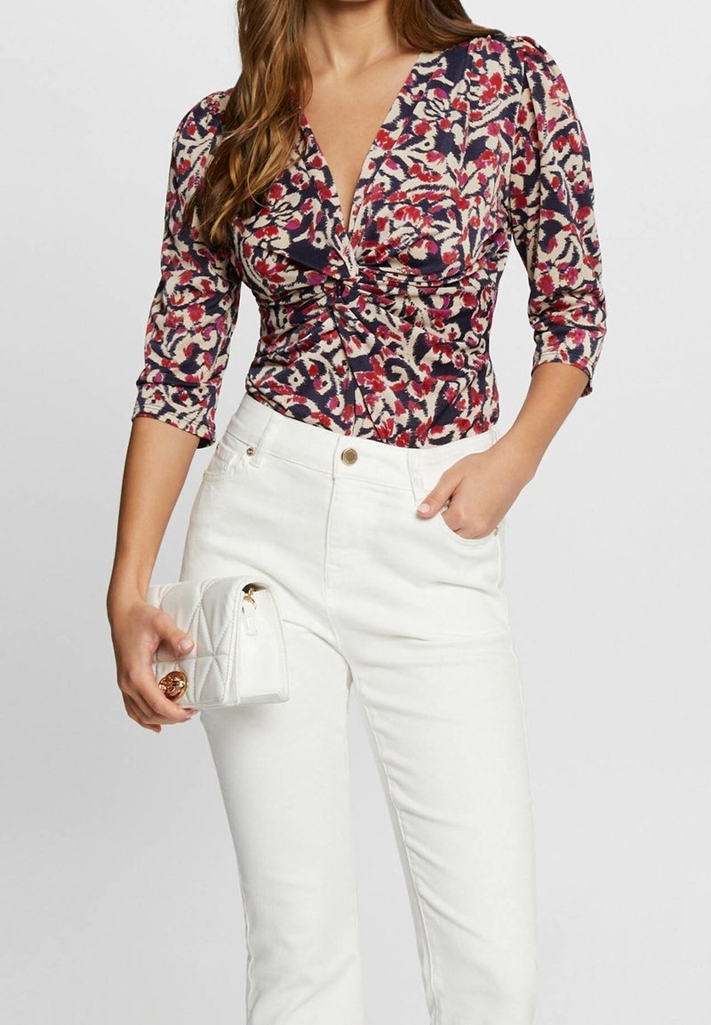 Blouse à motif floral en crème, marine et bordeaux avec des manches trois-quarts et un col en V. Associée à un jean blanc taille haute et une pochette blanche.