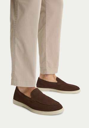 Braune Wildleder-Slipper mit weißen Sohlen, getragen zu hellbeigen Hosen, die Beine werden von der Mitte der Wade abwärts vor einem einfarbigen Hintergrund gezeigt.