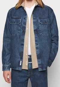 Chaqueta de mezclilla oscura con dos bolsillos en el pecho, cierre de botones y una camisa beige en contraste debajo. Modelo vistiendo jeans de mezclilla a juego.