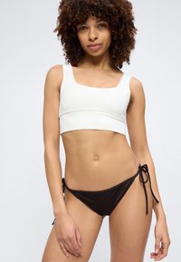 Jeune femme aux cheveux bouclés portant un haut court blanc et un bas de bikini noir à lanières latérales, debout devant un fond clair uni.