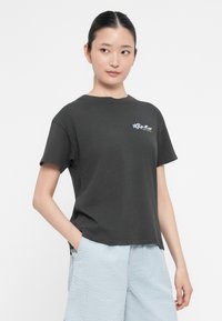 Črna bombažna majica s kratkimi rokavi, ki ima majhen modro cvetlični logotip na levem prsnem delu in sproščen kroj.