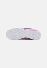 Lila és rózsaszín sneaker, texturált felsőrésszel és fehér gumitalppal, amely hullámos mintázatú. Nike logó látható a talpon.