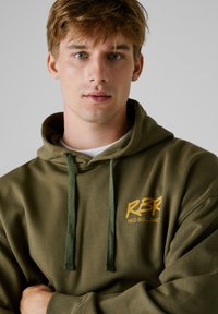 Hoodie vert olive avec cordons de serrage et un grand logo jaune affichant "RBR RED BULL RACE" en évidence sur la poitrine. Texture douce.